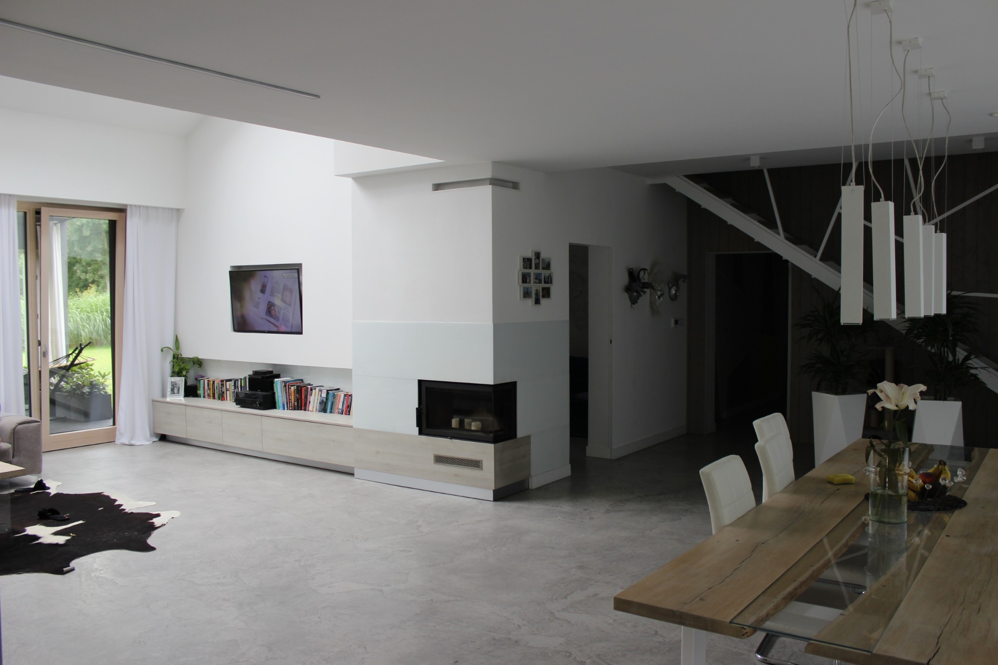 Przestronne, jasne wnętrze domu z kominkiem, telewizorem, drewnianym stołem z szklanym blatem i dywanem imitującym skórę krowy na szarej betonowej podłodze.