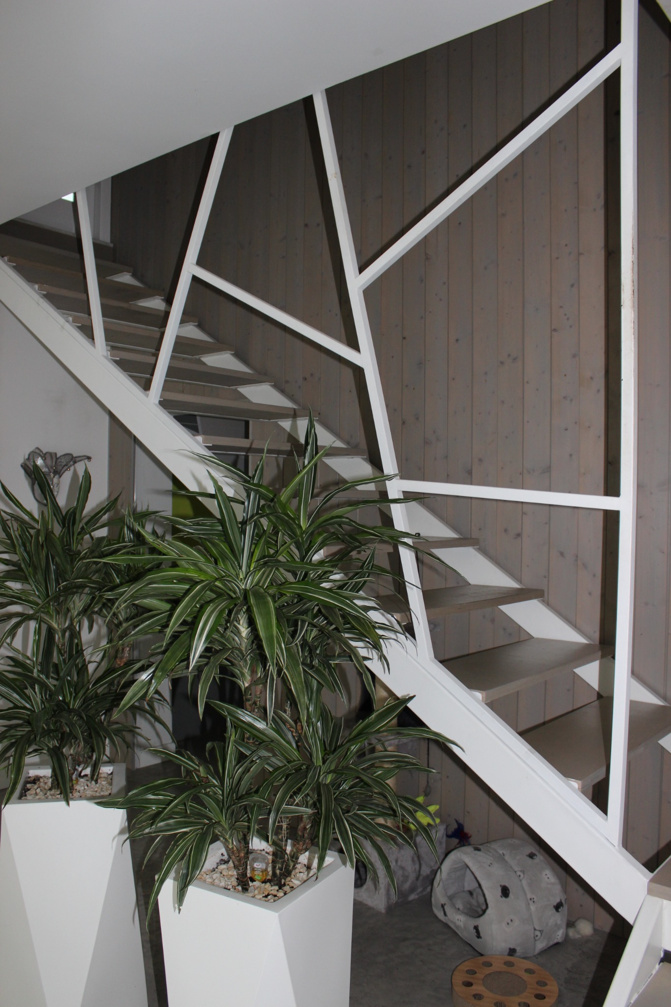 Nowoczesne schody z białymi, geometrycznymi barierkami, drewniane stopnie, ściana wyłożona pionowymi panelami z szarego drewna, donice z zielonymi roślinami.