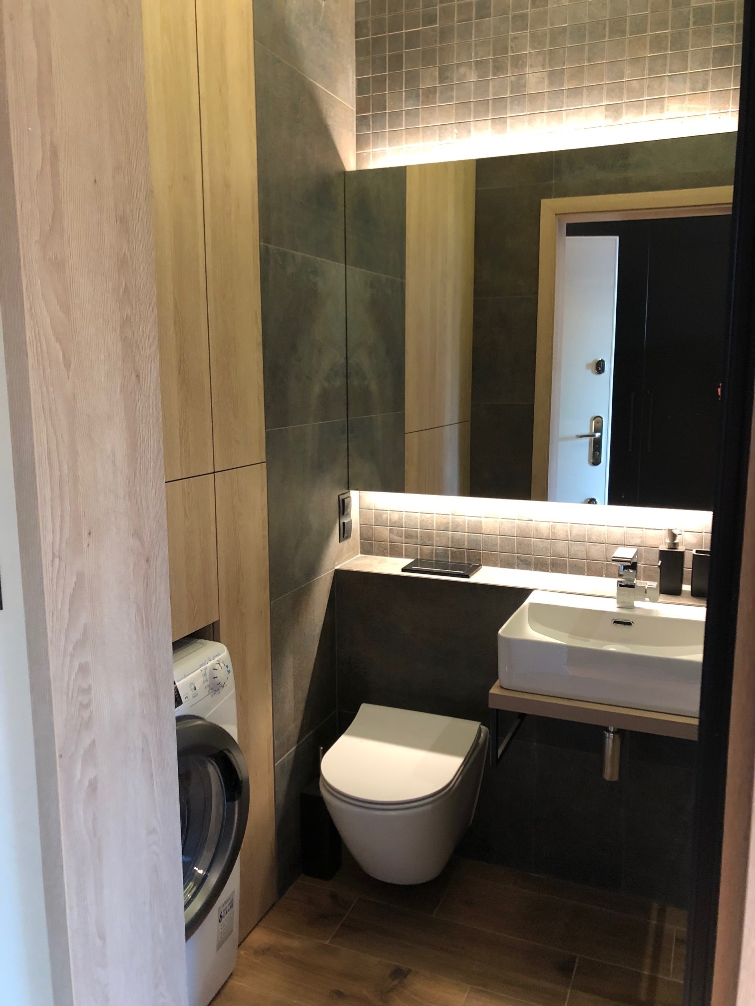 Nowoczesna łazienka z drewnianymi akcentami: szafka z pralką, umywalka podświetlona LED, toaleta i lustro na tle szarych płytek.
