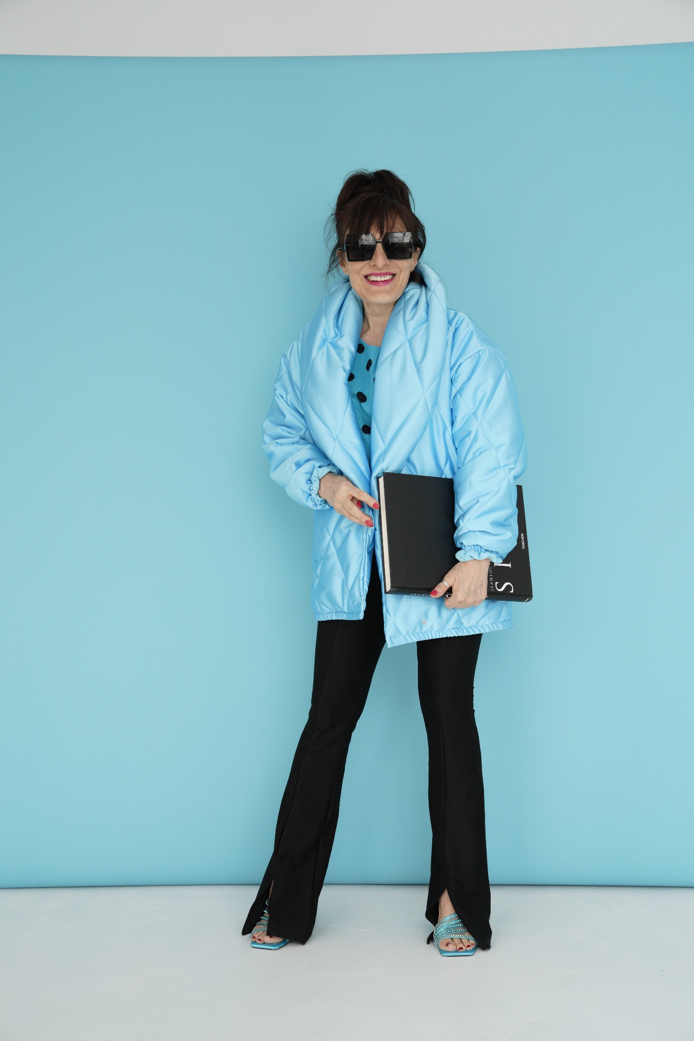 Uśmiechnięta kobieta w puchowej, błękitnej kurtce, czarnych spodniach z rozcięciem i turkusowych sandałach pozuje na tle błękitnej ściany, trzymając czarny album.