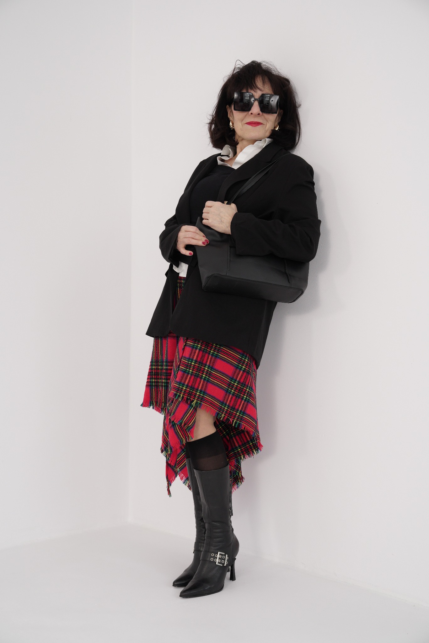 Kobieta w czarnej marynarce, spódnicy w szkocką kratę, podkolanówkach i skórzanych kozakach opiera się o białą ścianę w rogu, trzymając czarną torebkę.