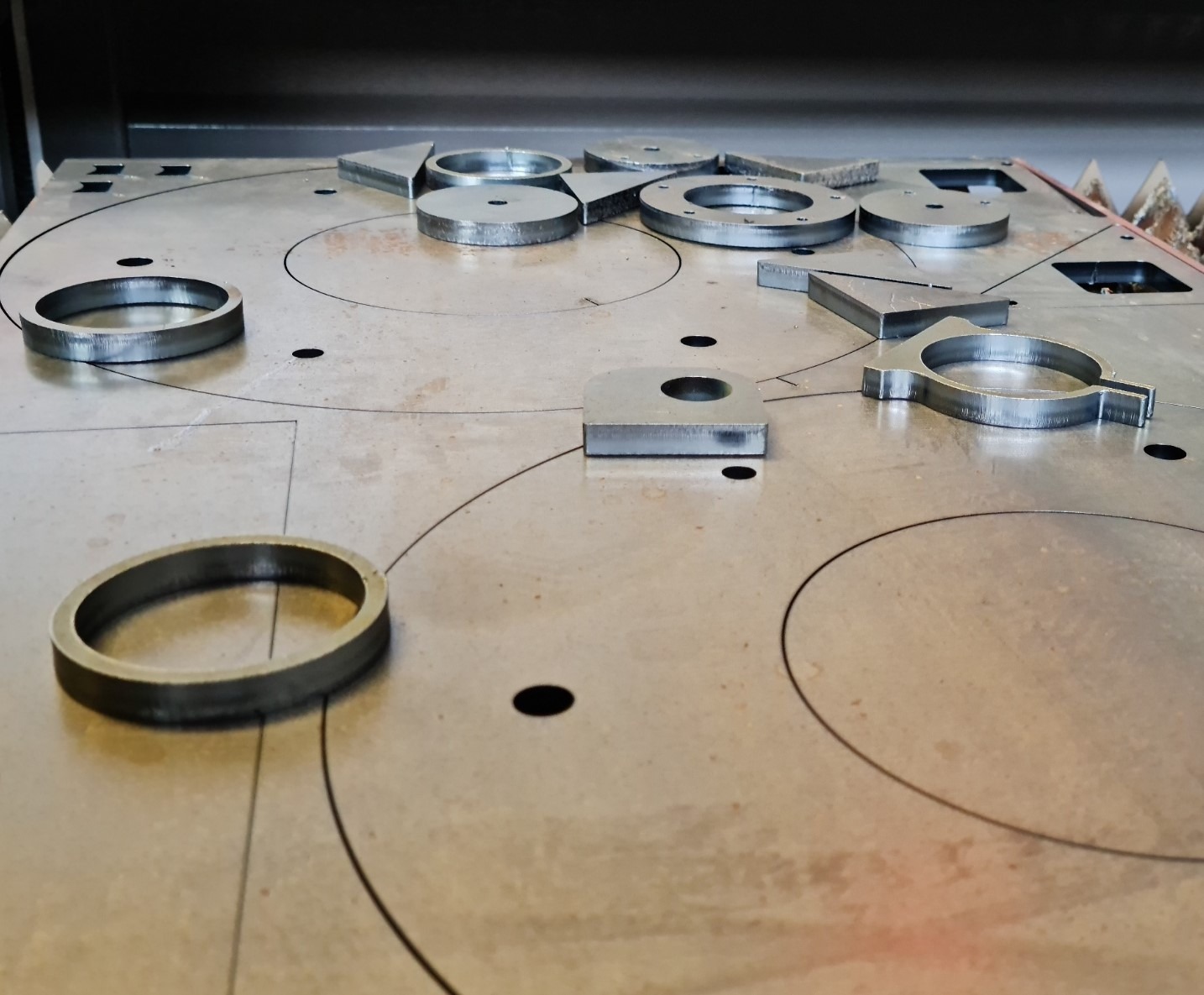 Metalowe elementy o różnych kształtach, wycięte z grubej blachy, leżące na metalowym stole z wygrawerowanymi liniami cięcia.