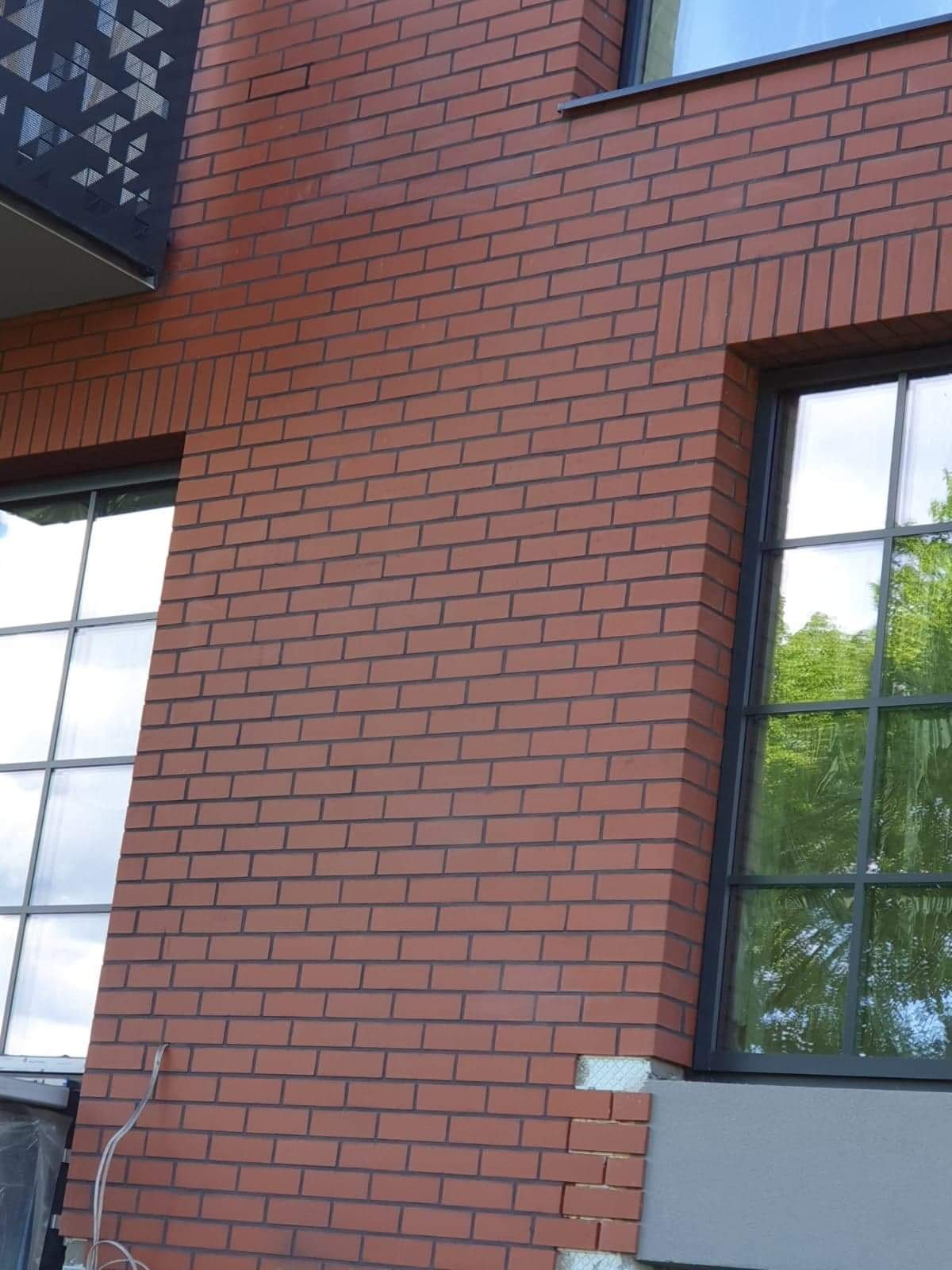 Fragment elewacji budynku z czerwonej cegły klinkierowej, widoczne okna z czarnymi ramami i fragment balkonu z ażurową balustradą, w dolnej części ściany widoczne niedokończone prace.
