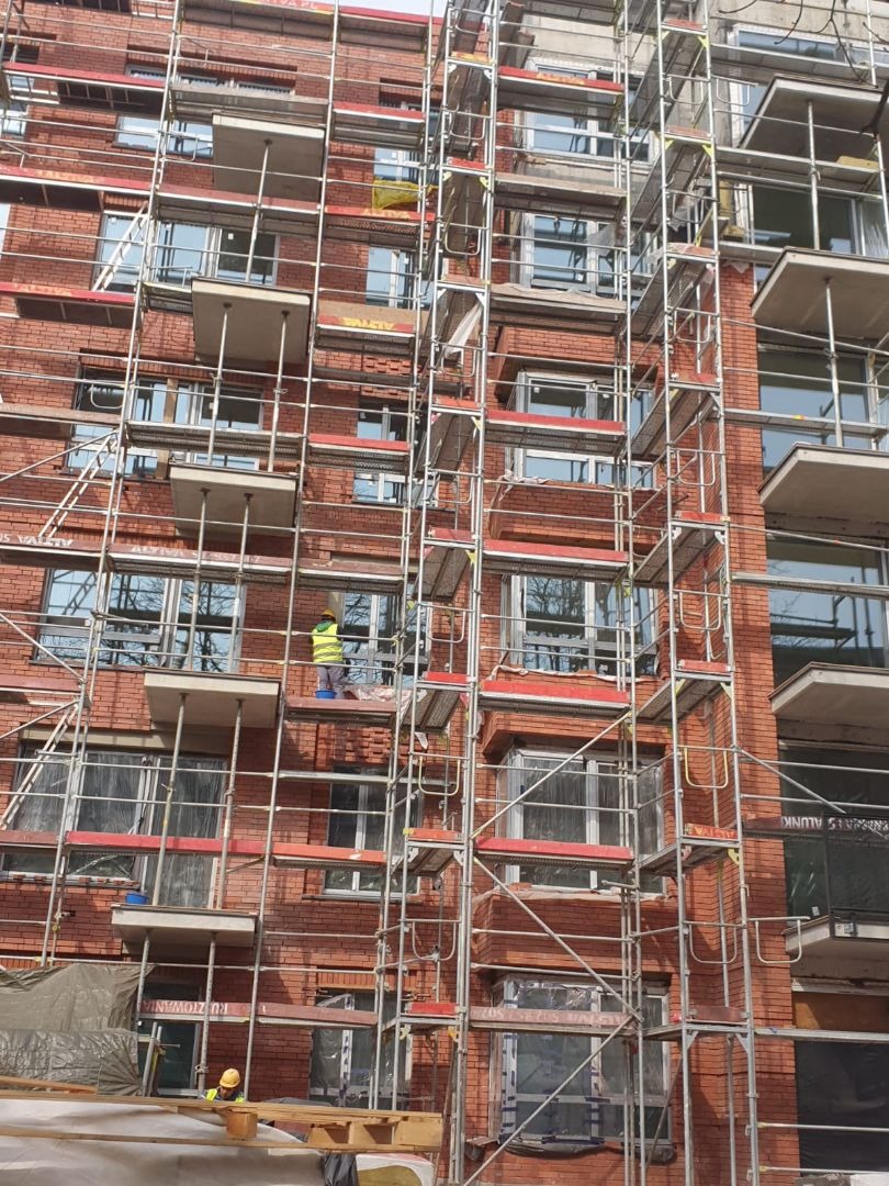 Murarz w żółtej kamizelce i kasku pracuje na rusztowaniu przy murowanej elewacji budynku z oknami, widoczne narzędzia i materiały budowlane.