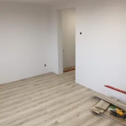 Jasne panele podłogowe ułożone w pokoju z białymi ścianami, widoczne narzędzia i pozostałości paneli przy ścianie.