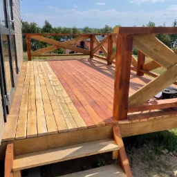 Nowo wybudowany drewniany taras z balustradą w kształcie krzyży, z częściowo pomalowaną podłogą w odcieniach czerwieni i naturalnego drewna, oraz schodami prowadzącymi do ogrodu.