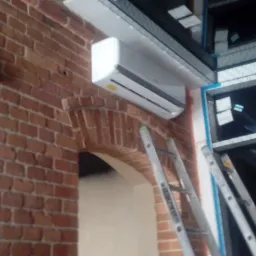 Biała jednostka klimatyzacyjna zamontowana na ceglanej ścianie z łukowym otworem, widoczna drabina aluminiowa.
