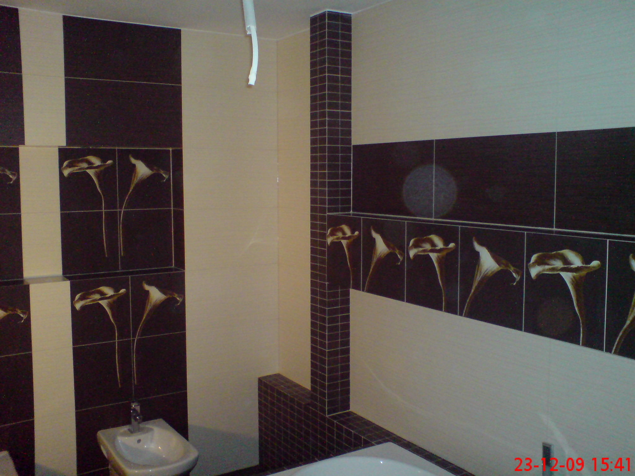 Wykończona łazienka z brązowo-beżowymi płytkami z motywem kalii, widoczna umywalka bidetowa i fragment wanny, brązowa kolumna z mozaiki.