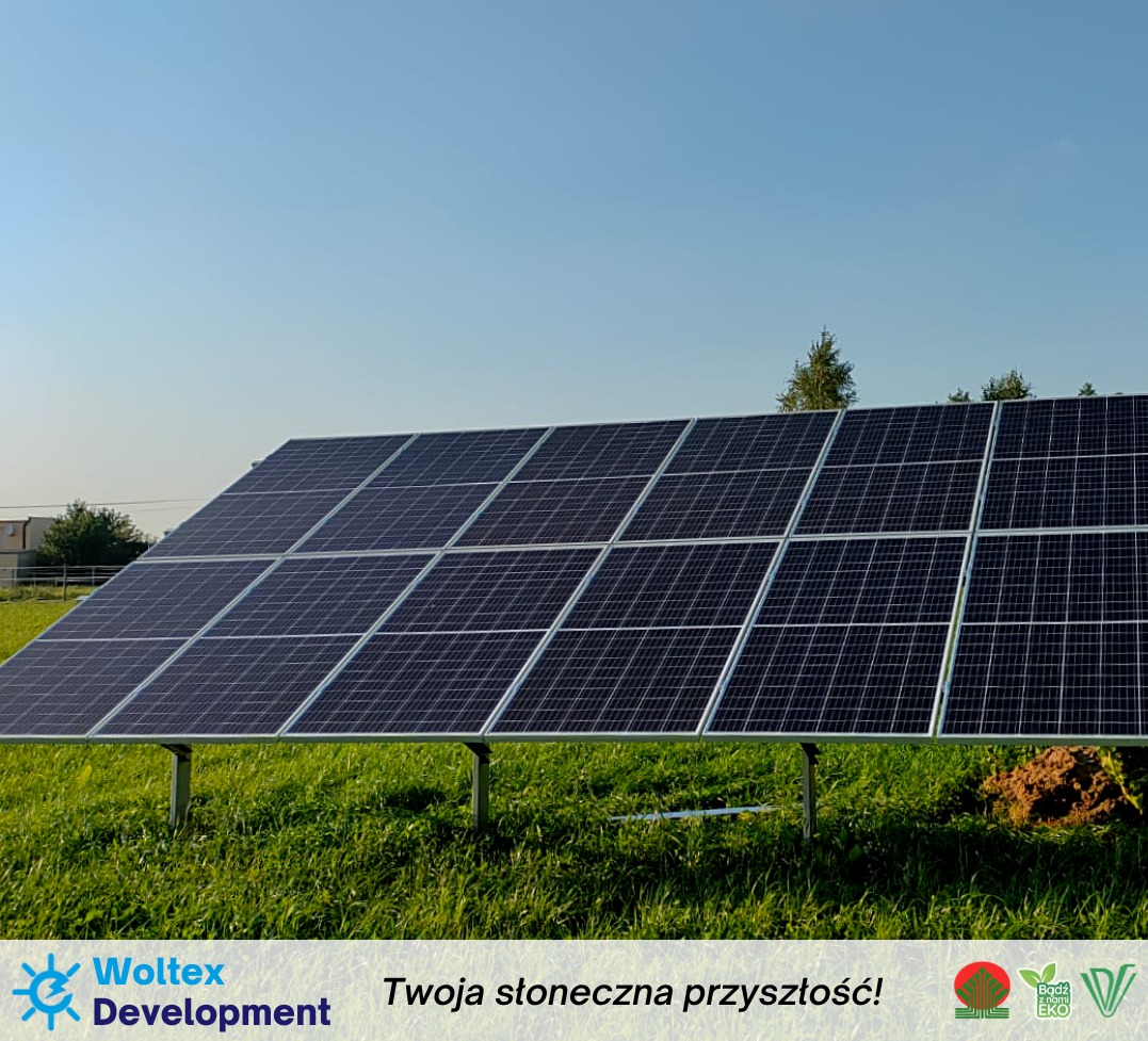 Panel słoneczny zainstalowany na metalowej konstrukcji na zielonej łące pod błękitnym niebem. Widoczne logo firmy Woltex Development w dolnej części zdjęcia.