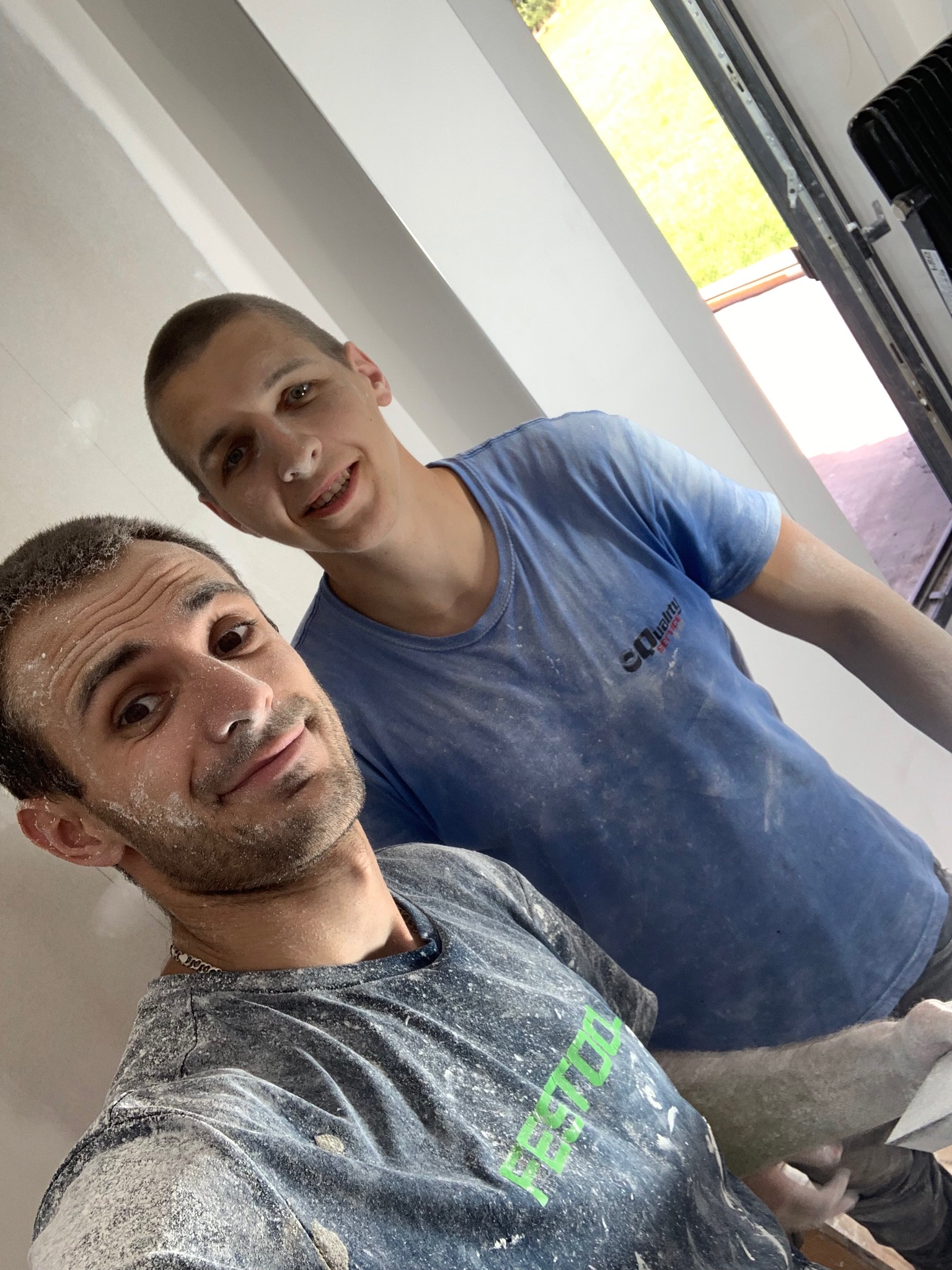 Dwóch uśmiechniętych mężczyzn, ubrudzonych gładzią szpachlową, pozuje do selfie na tle świeżo wyszpachlowanej ściany z oknem dachowym.