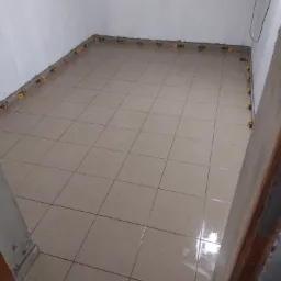 Ułożone beżowe płytki podłogowe w trakcie montażu, z żółtymi klipsami poziomującymi przy ścianach, w pomieszczeniu z białymi ścianami.