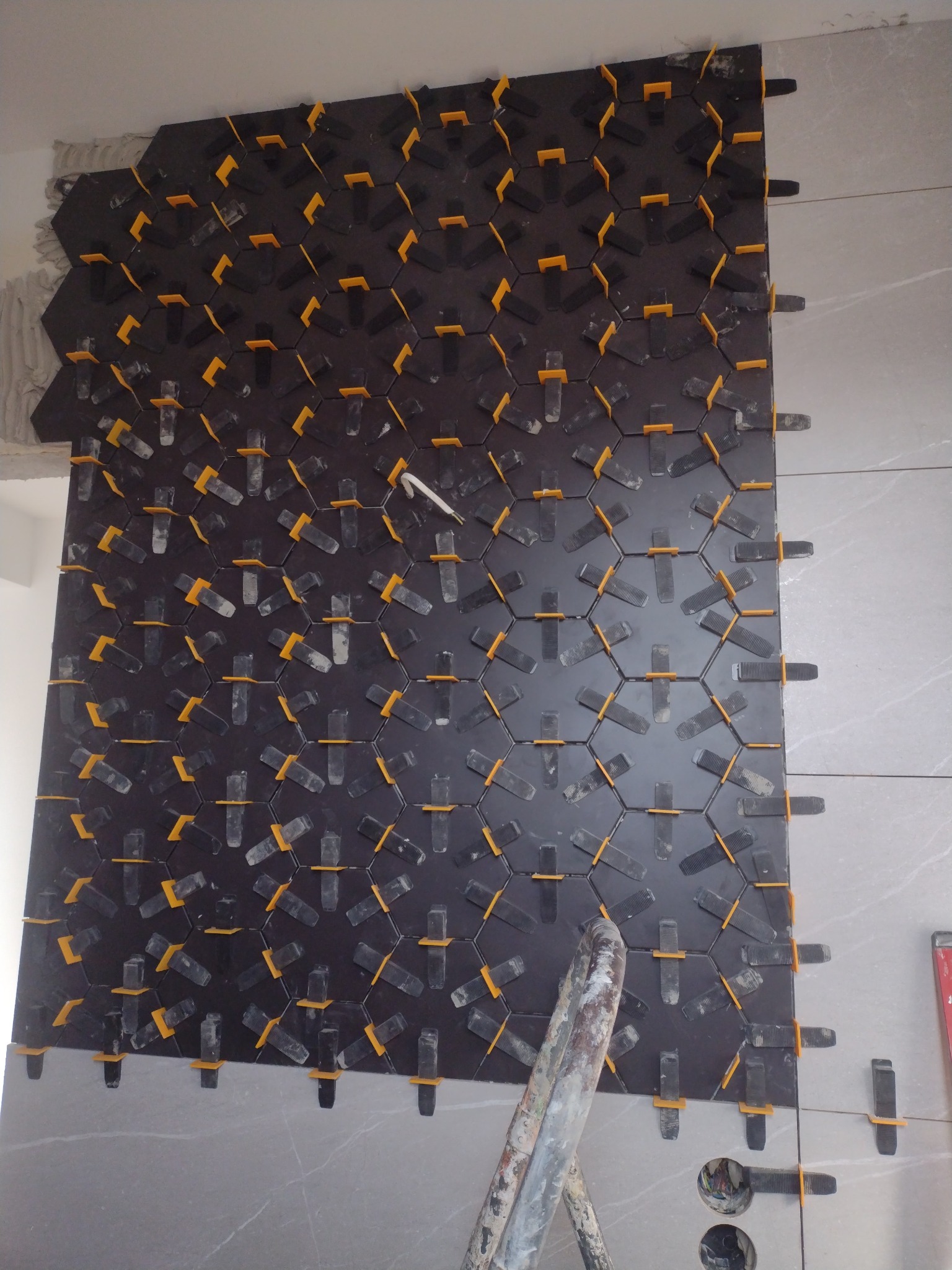 Układanie heksagonalnych, czarnych płytek na ścianie z użyciem żółtych klipsów poziomujących, widoczne niedociągnięcia przy suficie i rurka instalacyjna na dole.