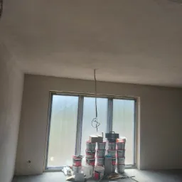 Surowe wnętrze po gruntowaniu ścian i sufitu, z oknem zabezpieczonym folią i stertą puszek z farbą przy nim, przewody elektryczne zwisające z sufitu.