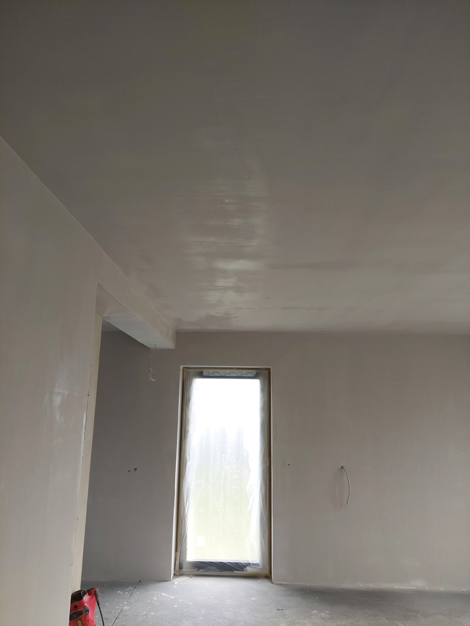 Świeżo wykończone wnętrze z oknem zabezpieczonym folią, ściany i sufit po gruntowaniu, widoczne ślady szpachlowania, na podłodze resztki materiałów budowlanych, w rogu pomieszczenia czerwony...