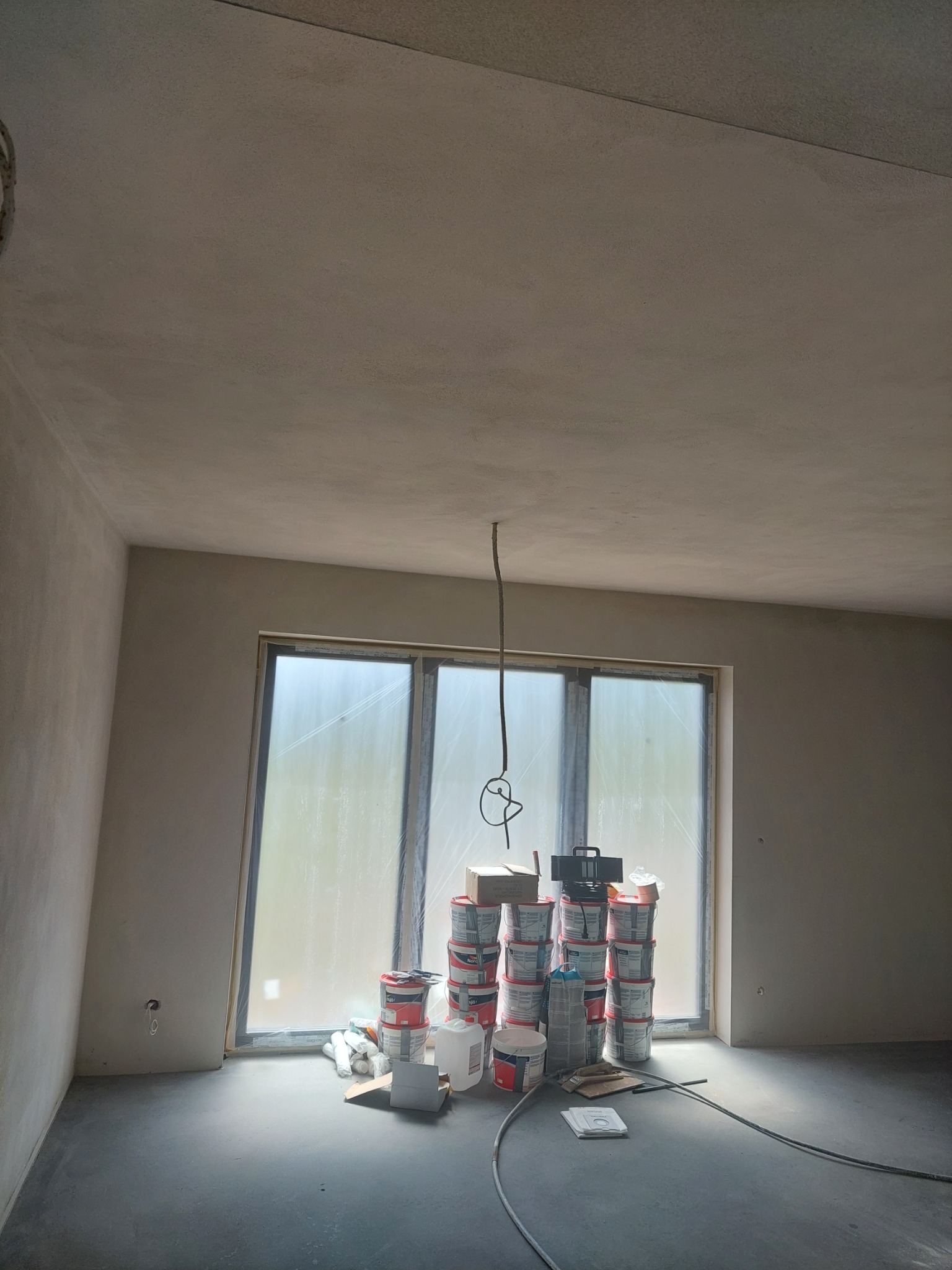 Surowe wnętrze po gruntowaniu ścian i sufitu, z oknem zabezpieczonym folią i stertą puszek z farbą przy nim, przewody elektryczne zwisające z sufitu.