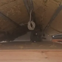 Izolacja poddasza wełną mineralną, widoczne metalowe profile, zwisający kabel elektryczny i fragment okna w tle.