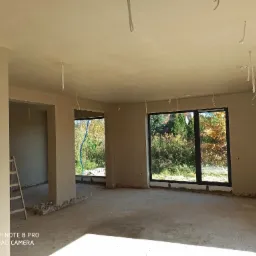 Surowe wnętrze po nałożeniu tynków maszynowych, widoczne kable zwisające z sufitu, ślady po pracach na podłodze, drabina oparta o ścianę, duże okna z widokiem na zieleń.