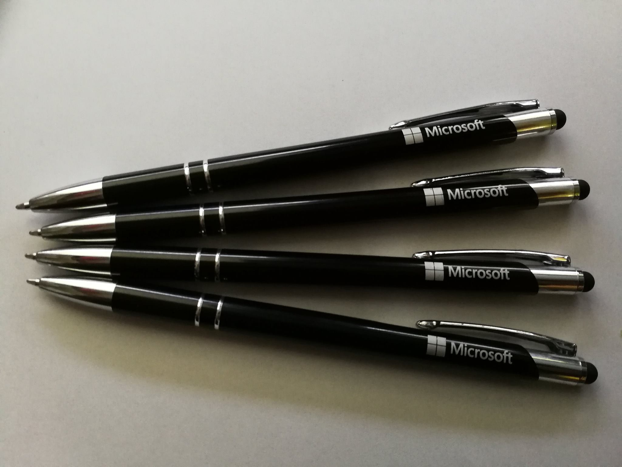Cztery czarne długopisy z logo Microsoft ułożone równolegle na jasnym tle, każdy z metalowym klipsem i gumką dotykową na końcu.