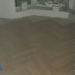 Układanie parkietu w jodełkę: widoczne fragmenty ułożonej podłogi, białe ściany, narzędzia i puszki po materiałach budowlanych przy ścianie i niebieska taśma malarska przy podłodze.