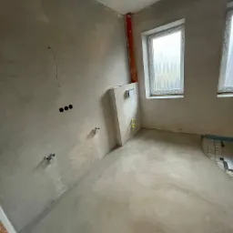 Surowa łazienka w trakcie prac przygotowawczych: ściany pokryte szpachlą, widoczne instalacje wodne, przygotowane podłoże pod ogrzewanie podłogowe.