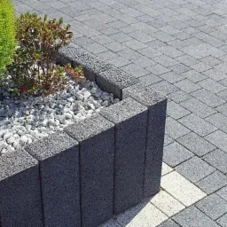 Krawężnik z ciemnych, pionowych kostek obramowujący rabatę z białym grysem i roślinami, przylegający do szarej nawierzchni z kostki brukowej z jasnymi akcentami.