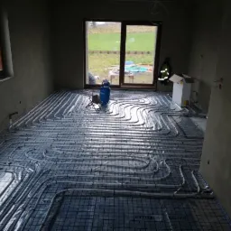 Wnętrze w trakcie montażu ogrzewania podłogowego: rury grzewcze ułożone na siatce, widoczna butla gazowa, surowe ściany i okna z widokiem na zewnątrz.