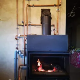 Otwarty kominek z palącym się drewnem, obudowany instalacją z miedzianych rur grzewczych, manometr, szara ściana w tle.