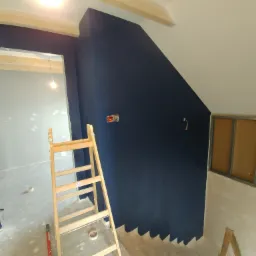 Wnętrze w trakcie remontu: ściana pomalowana na granatowo, widoczne schody, drabina, otwór drzwiowy i fragment konstrukcji pod płytę karton gips.