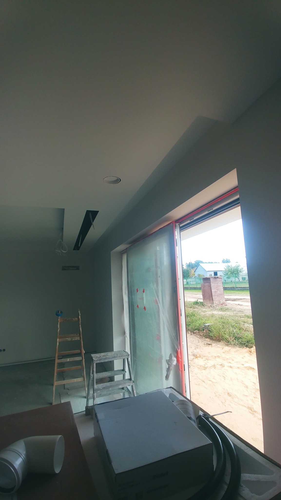 Wnętrze w trakcie prac wykończeniowych w Rzeszowie: widoczne drabiny, okno zabezpieczone folią, sufit z przygotowanymi otworami na oświetlenie, elementy instalacji wentylacyjnej i materiały budowlane.