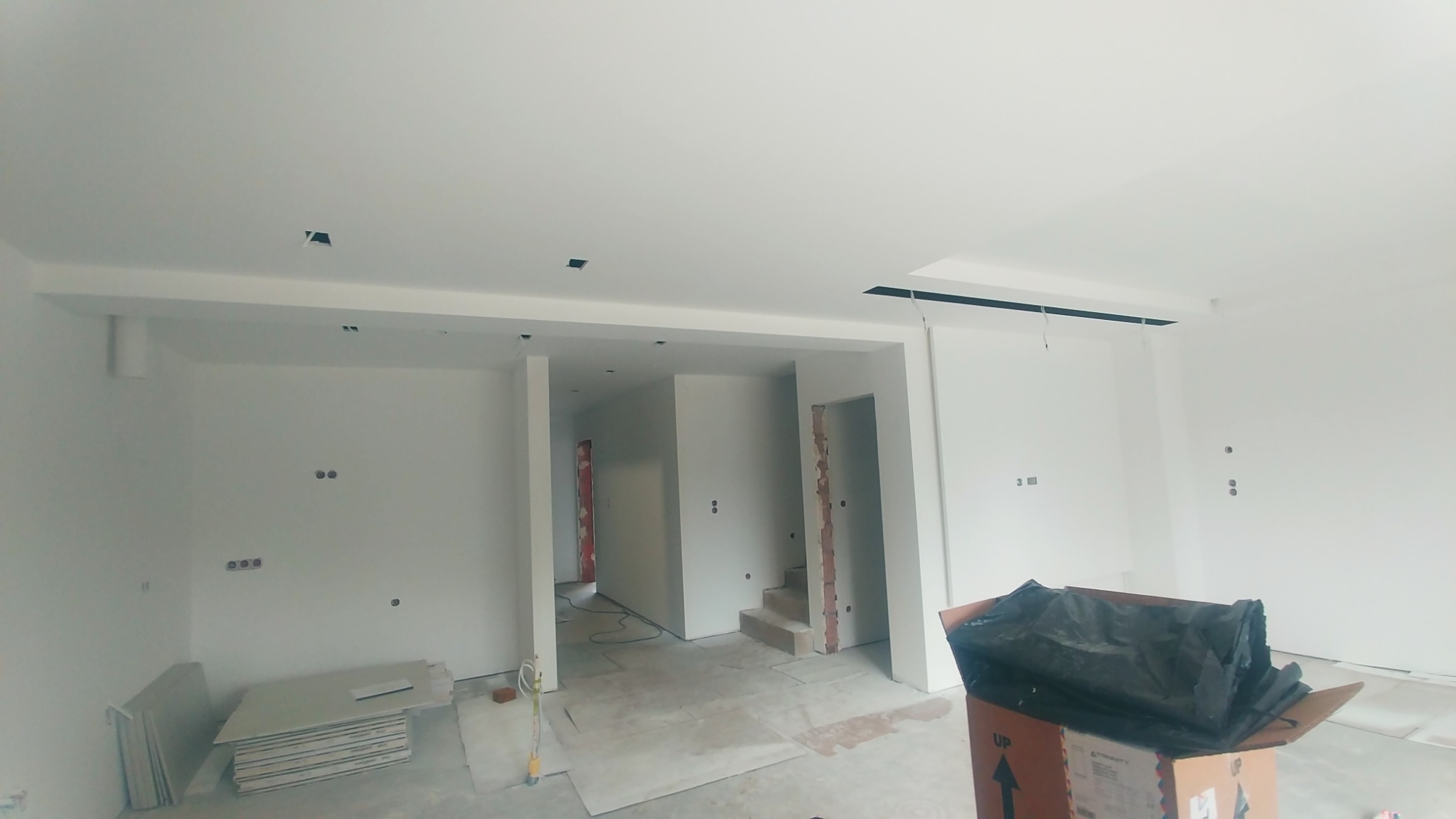 Wnętrze w trakcie prac wykończeniowych: świeżo pomalowane ściany na biało, widoczne puszki elektryczne, przygotowane płyty karton-gips, karton z workiem na gruz, surowe schody w tle.