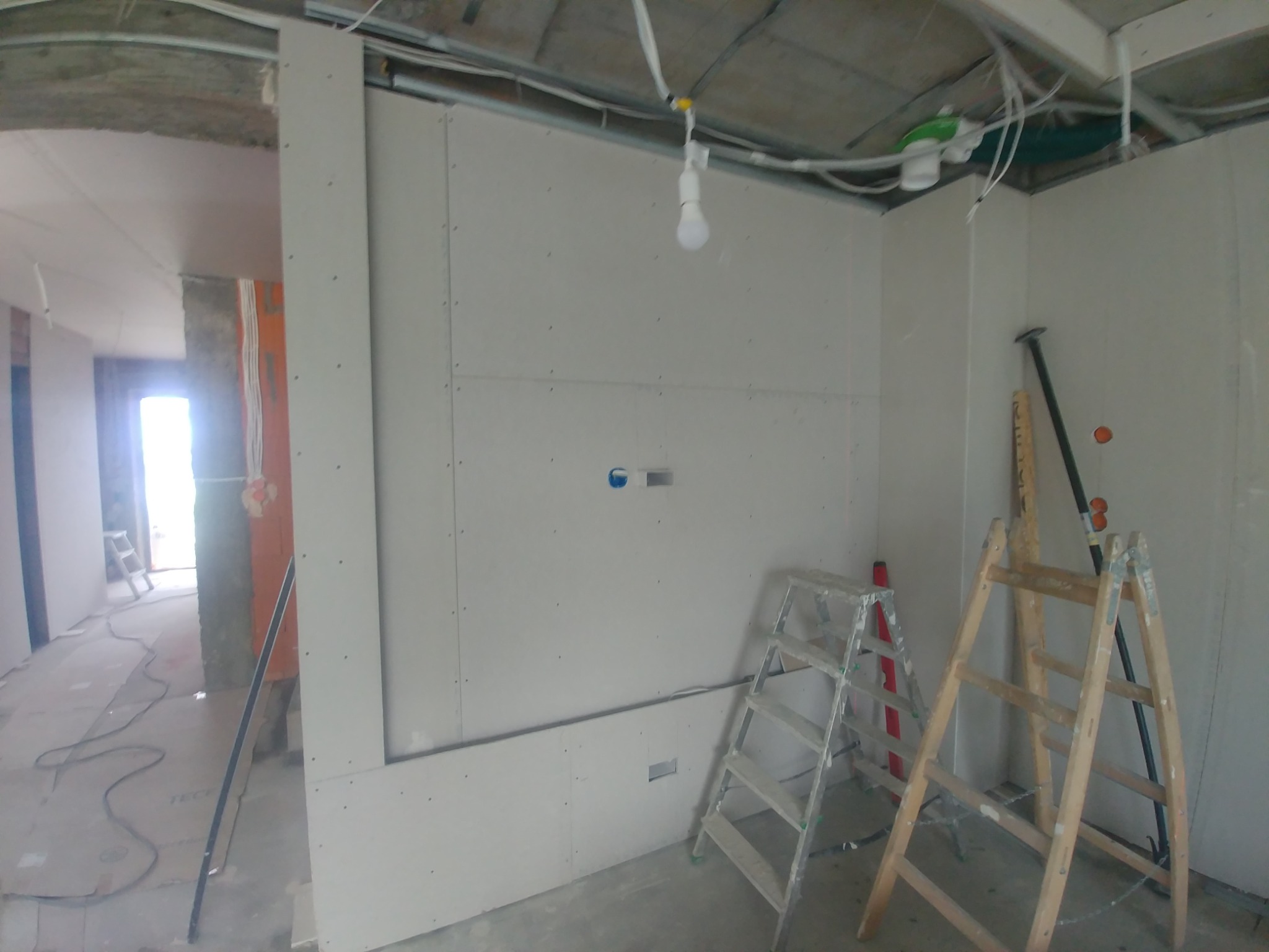 Wnętrze w trakcie remontu: ściany wyłożone płytami gipsowo-kartonowymi, widoczne drabiny, przewody elektryczne pod sufitem i otwór drzwiowy do kolejnego pomieszczenia.