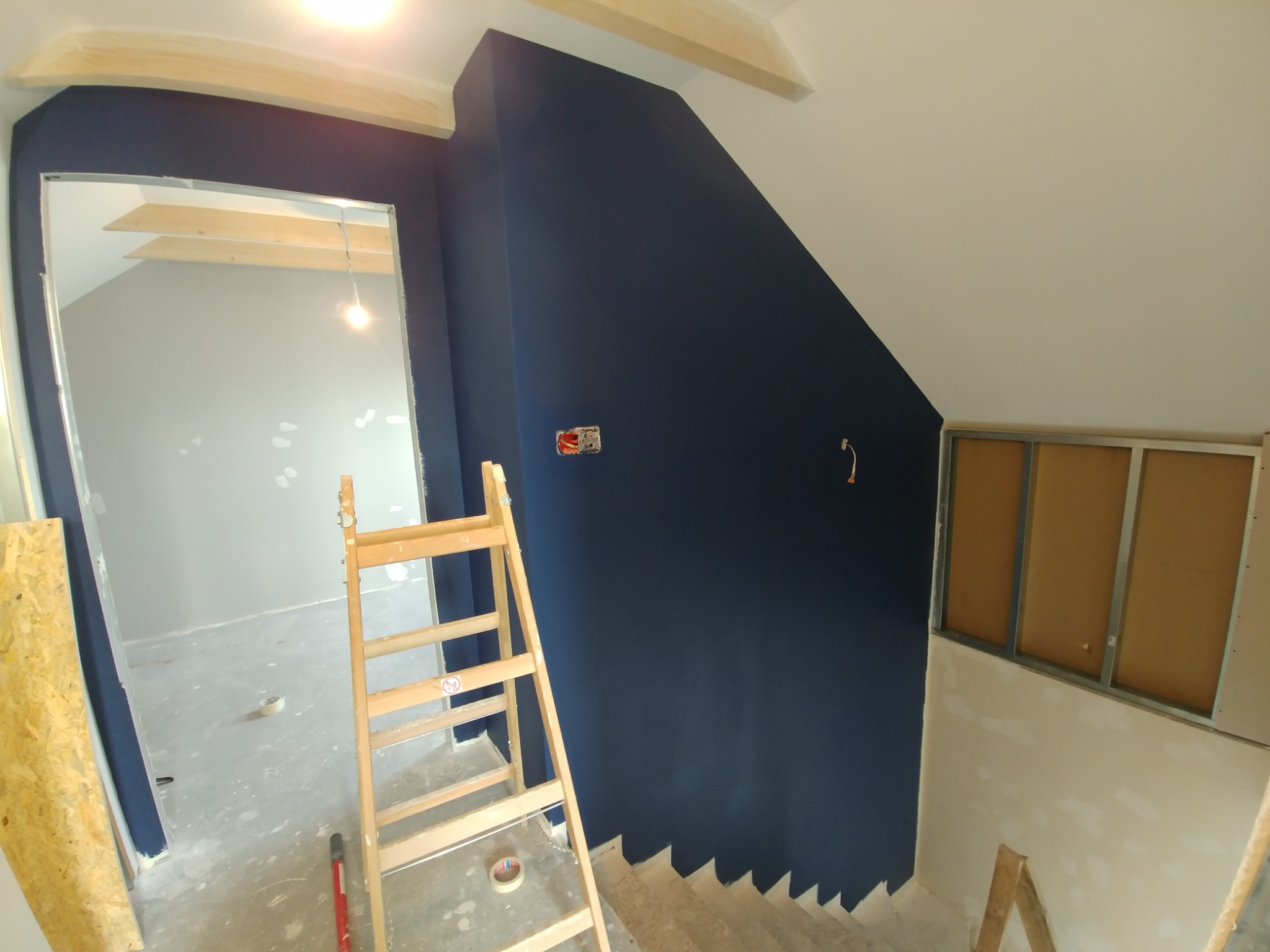 Wnętrze w trakcie remontu: ściana pomalowana na granatowo, widoczne schody, drabina, otwór drzwiowy i fragment konstrukcji pod płytę karton gips.