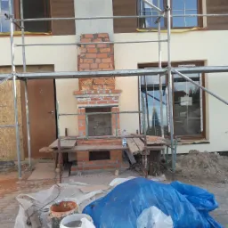 Wznoszenie kominka z cegły na zewnątrz budynku w trakcie budowy, widoczne rusztowanie, okna z brązowymi ramami, oraz materiały budowlane zabezpieczone folią.