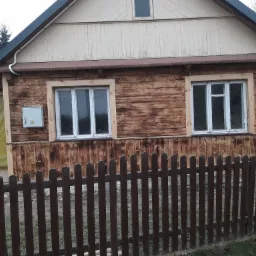 Drewniany dom z dwoma oknami w białych ramach, częściowo osłonięty brązowym płotem sztachetowym; widoczna skrzynka elektryczna na elewacji.