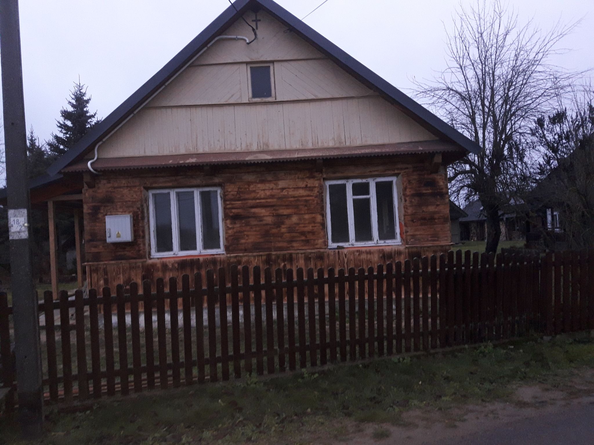 Drewniany dom z brązową elewacją i białymi oknami, ogrodzony niskim drewnianym płotem, widziany w pochmurny dzień.