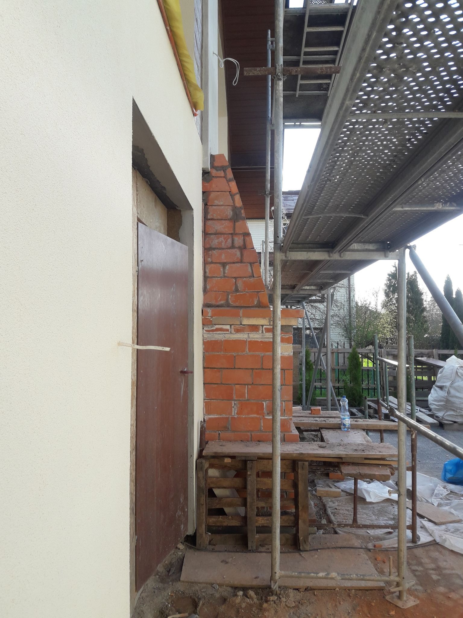 Budowa komina z czerwonej cegły przy ścianie budynku z zamontowanymi rusztowaniami, widoczna butelka wody na drewnianej konstrukcji.