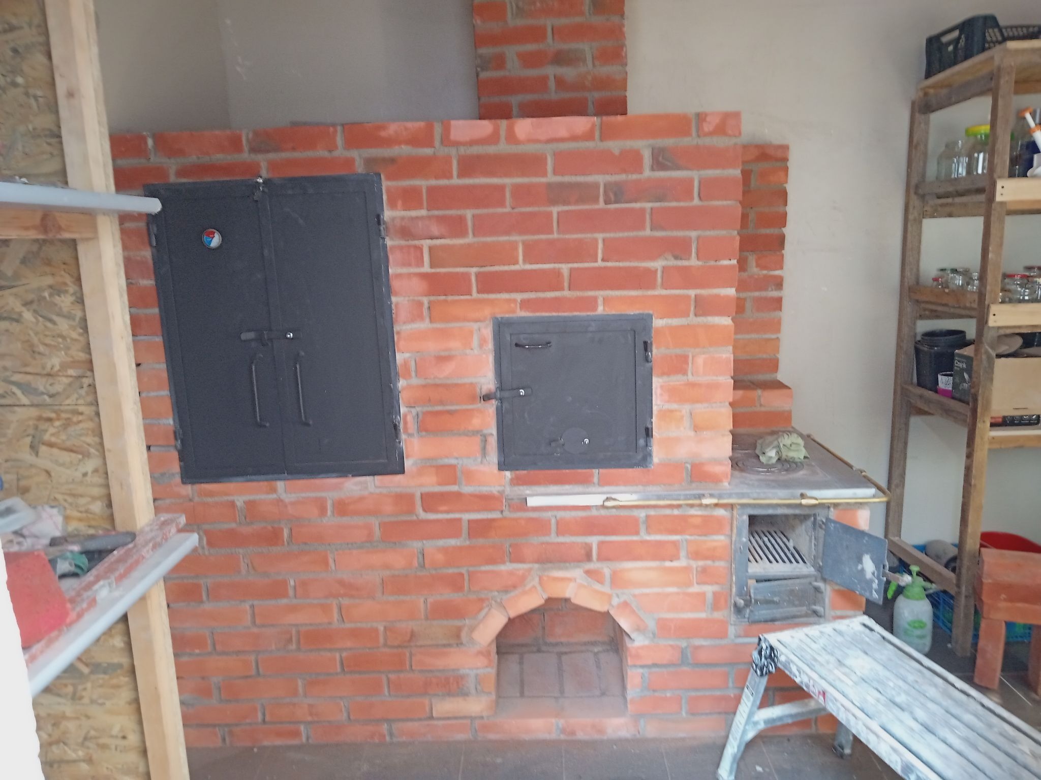 Murowany piec z czerwonej cegły z metalowymi drzwiczkami, widoczny fragment komina, drabina malarska i regał z słoikami w tle.