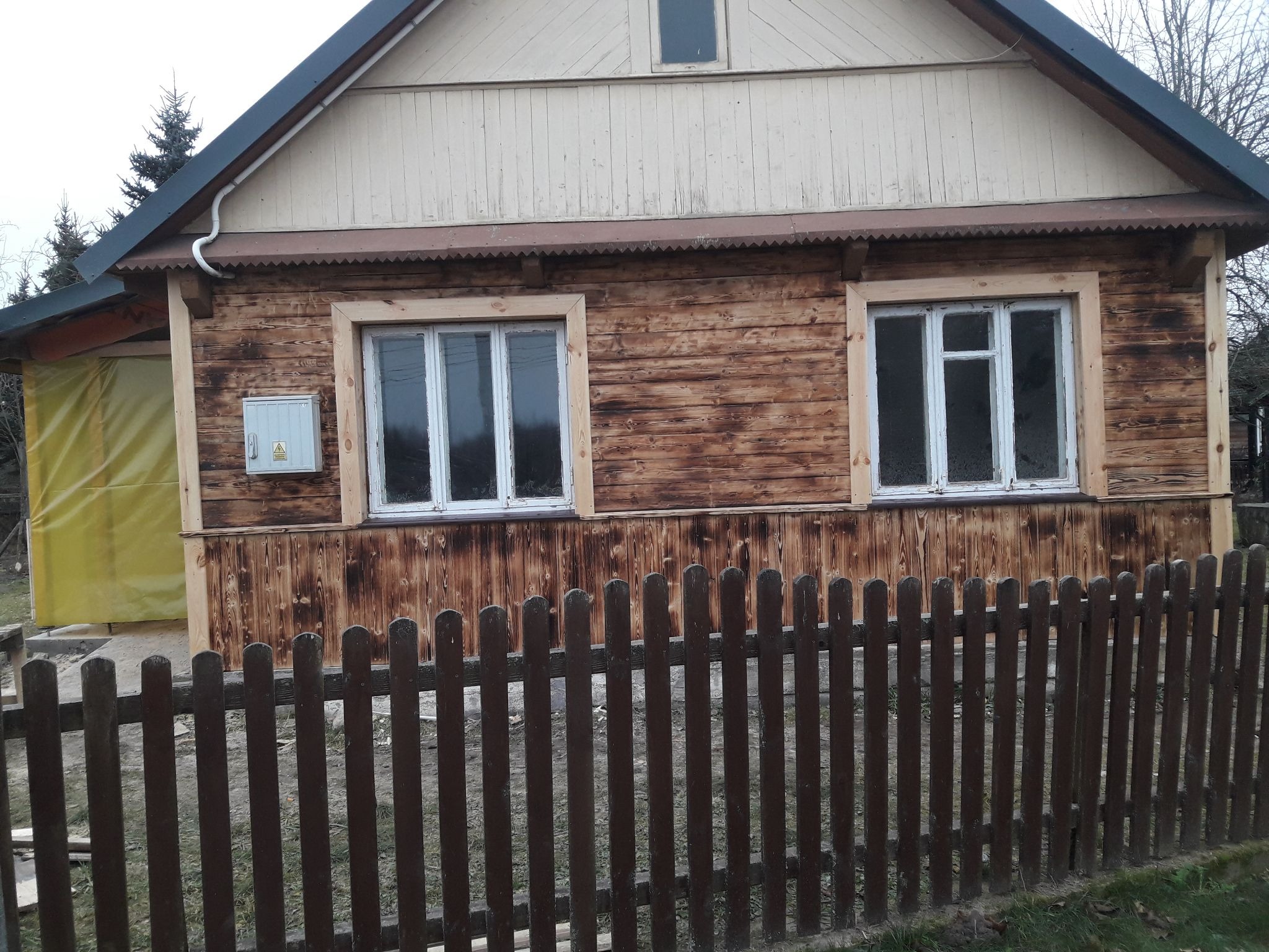 Drewniany dom z dwoma oknami w białych ramach, częściowo osłonięty brązowym płotem sztachetowym; widoczna skrzynka elektryczna na elewacji.