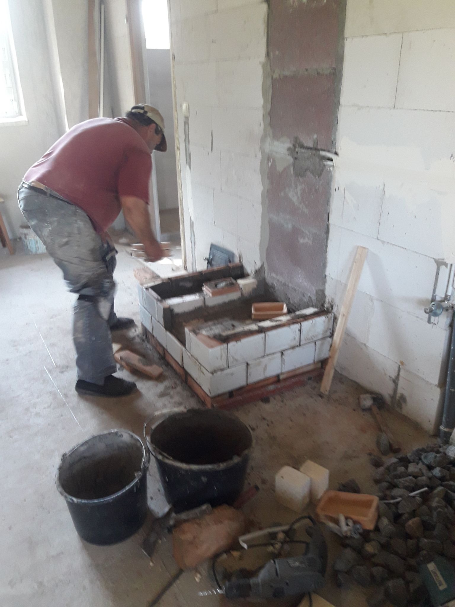 Budowa kominka z białych i czerwonych cegieł w surowym wnętrzu, widoczne narzędzia i materiały budowlane, w tle murarz w czerwonej koszulce.