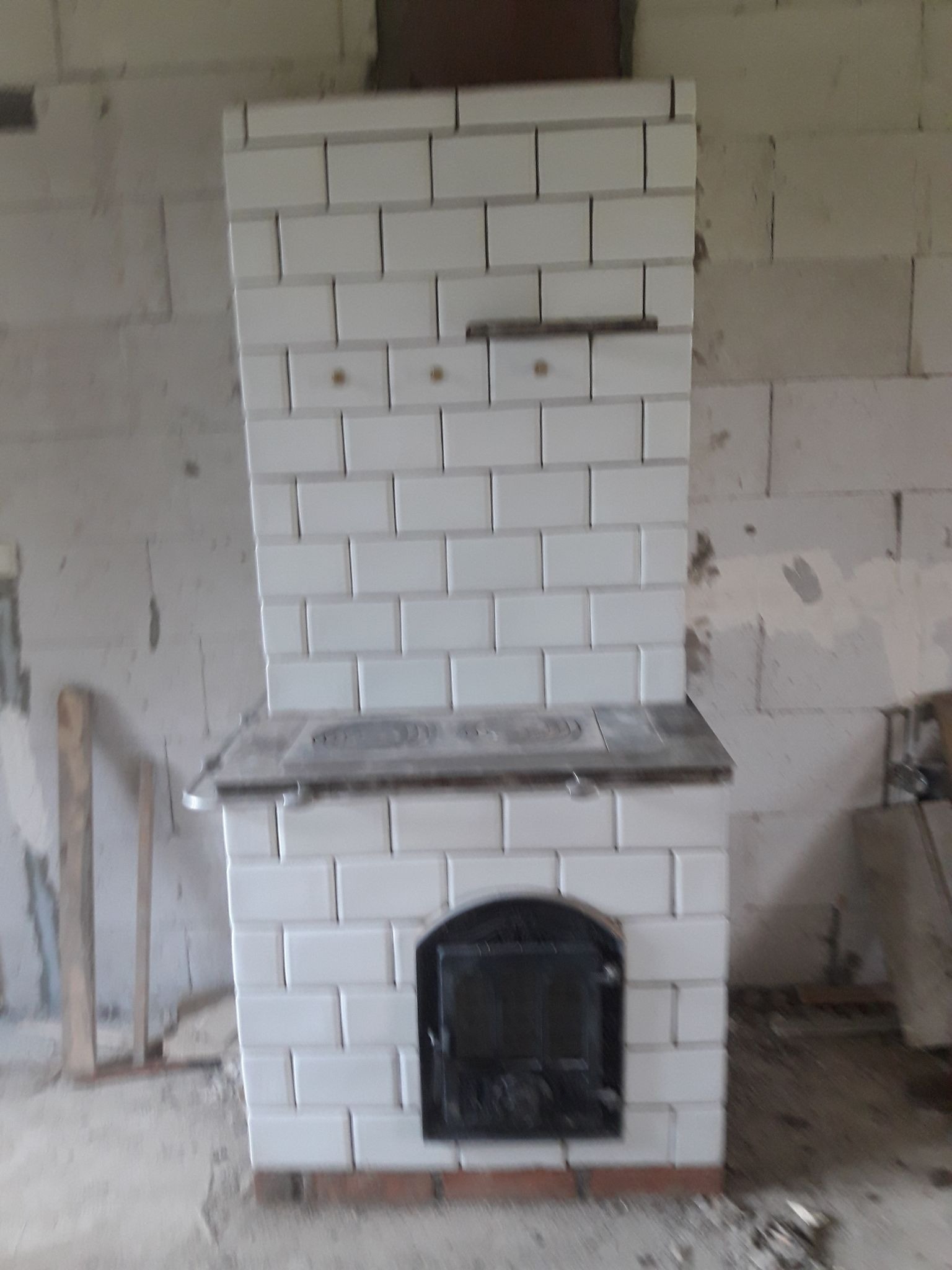 Biały piec kaflowy z czarnymi drzwiczkami w surowym wnętrzu, widoczne niedokończone ściany z bloczków.