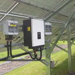 Rząd paneli słonecznych zamontowanych na metalowej konstrukcji na trawniku, widoczny inwerter i licznik energii.