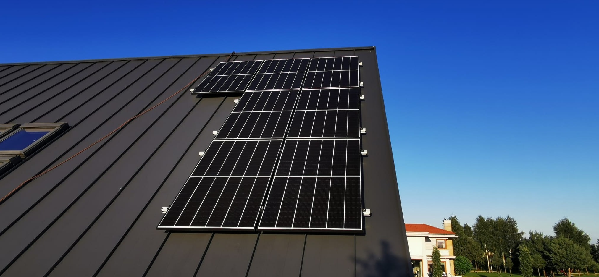 Częściowy widok instalacji paneli na dachu z blachy trapezowej, błękitne niebo w tle i fragment domu sąsiada.