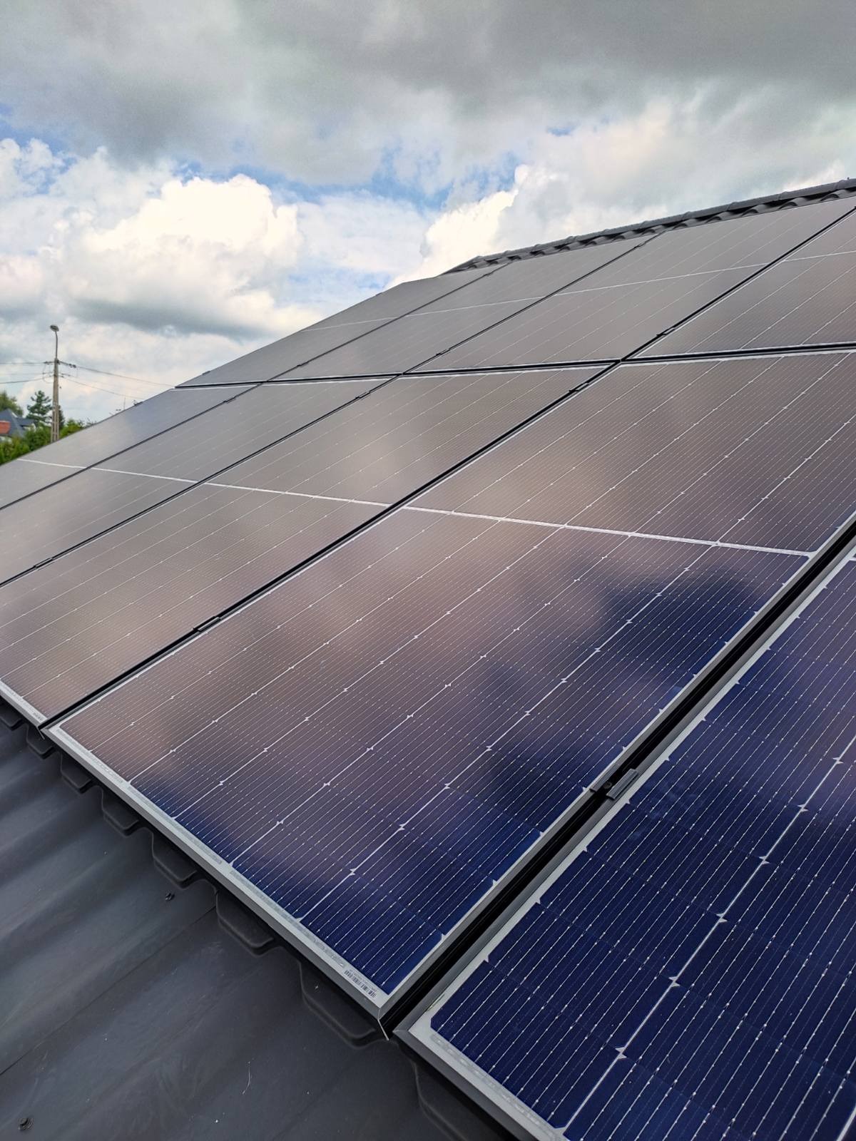 Ciemnoniebieskie panele fotowoltaiczne na ciemnoszarym dachu, odbijające chmury i błękit nieba.