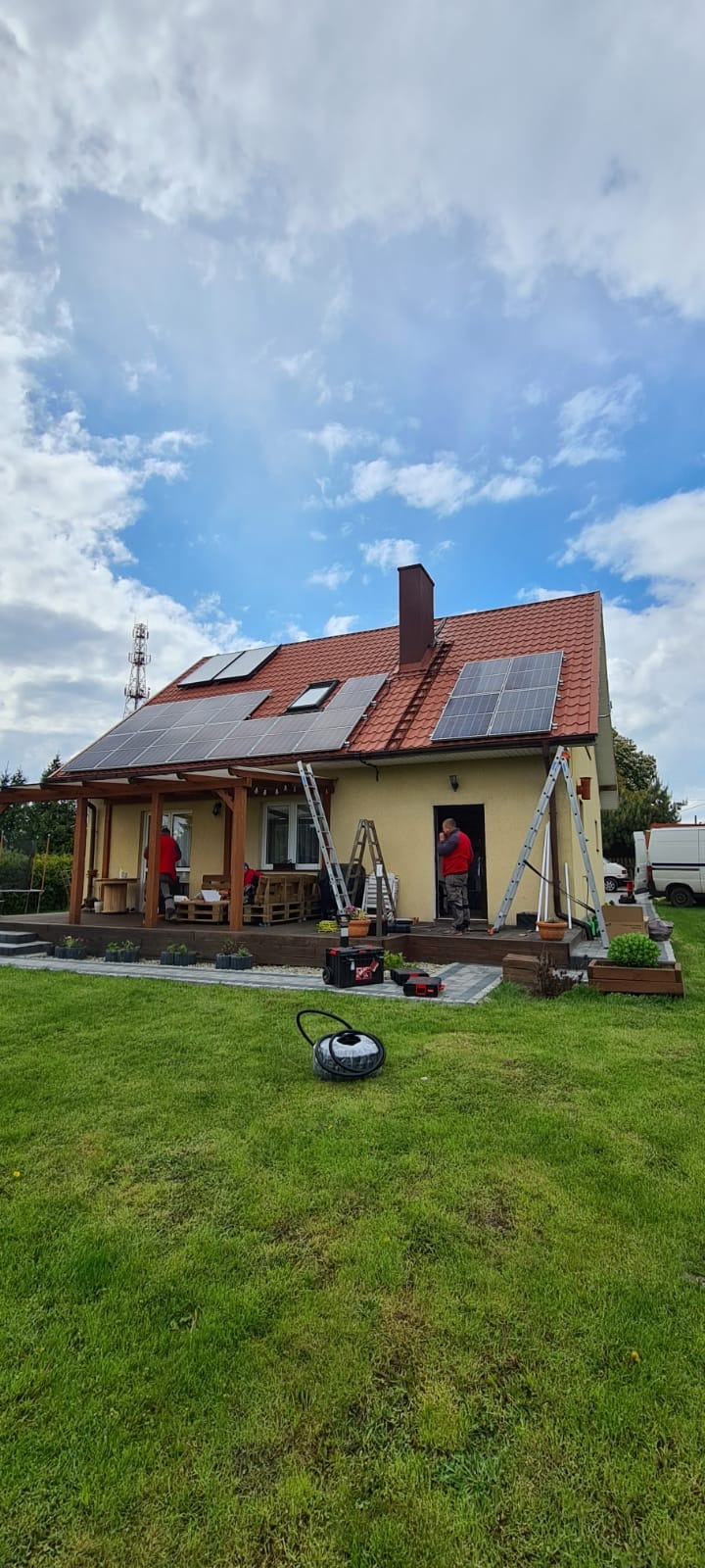 Montaż paneli na dachu domu jednorodzinnego z czerwonej dachówki, widoczne drabiny i dwóch monterów w czerwonych kamizelkach, na trawniku leży odkurzacz przemysłowy.
