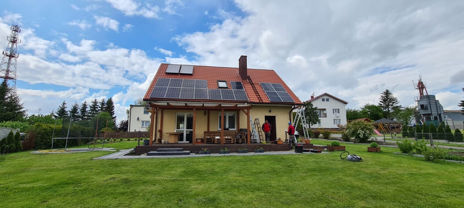 Montaż paneli słonecznych na dachu domu jednorodzinnego z czerwonej dachówki, widoczni dwaj monterzy na drabinach, w tle zielony trawnik i elementy infrastruktury telekomunikacyjnej.