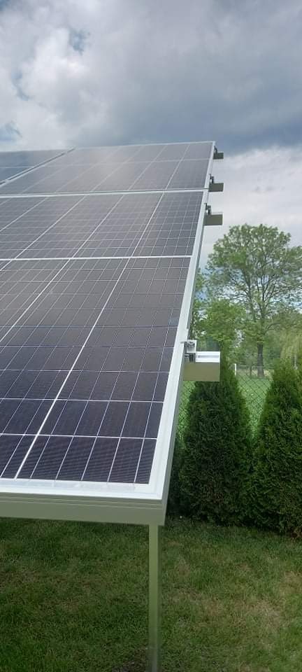 Panel słoneczny na aluminiowej konstrukcji wsporczej, widok z dołu na tle zieleni i pochmurnego nieba.