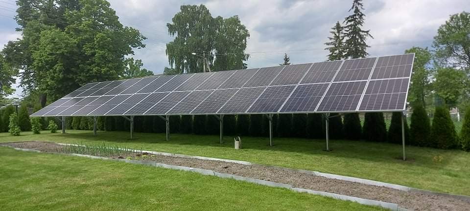 Duży panel słoneczny na metalowych podporach, zainstalowany na trawniku obok warzywnika z metalowym obramowaniem, drzewa w tle.