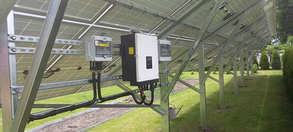 Rząd paneli słonecznych zamontowanych na metalowej konstrukcji na trawniku, widoczny inwerter i licznik energii.