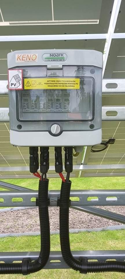 Szara skrzynka przyłączeniowa fotowoltaiki KENO Noark z naklejkami ostrzegawczymi, zamontowana na konstrukcji wsporczej paneli słonecznych, z widocznymi kablami w czarnych peszlach.