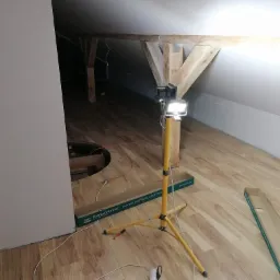 Poddasze z drewnianą konstrukcją więźby dachowej, oświetlone halogenem na statywie, z częściowo ułożoną podłogą z paneli drewnopodobnych i porozrzucanymi opakowaniami po panelach.